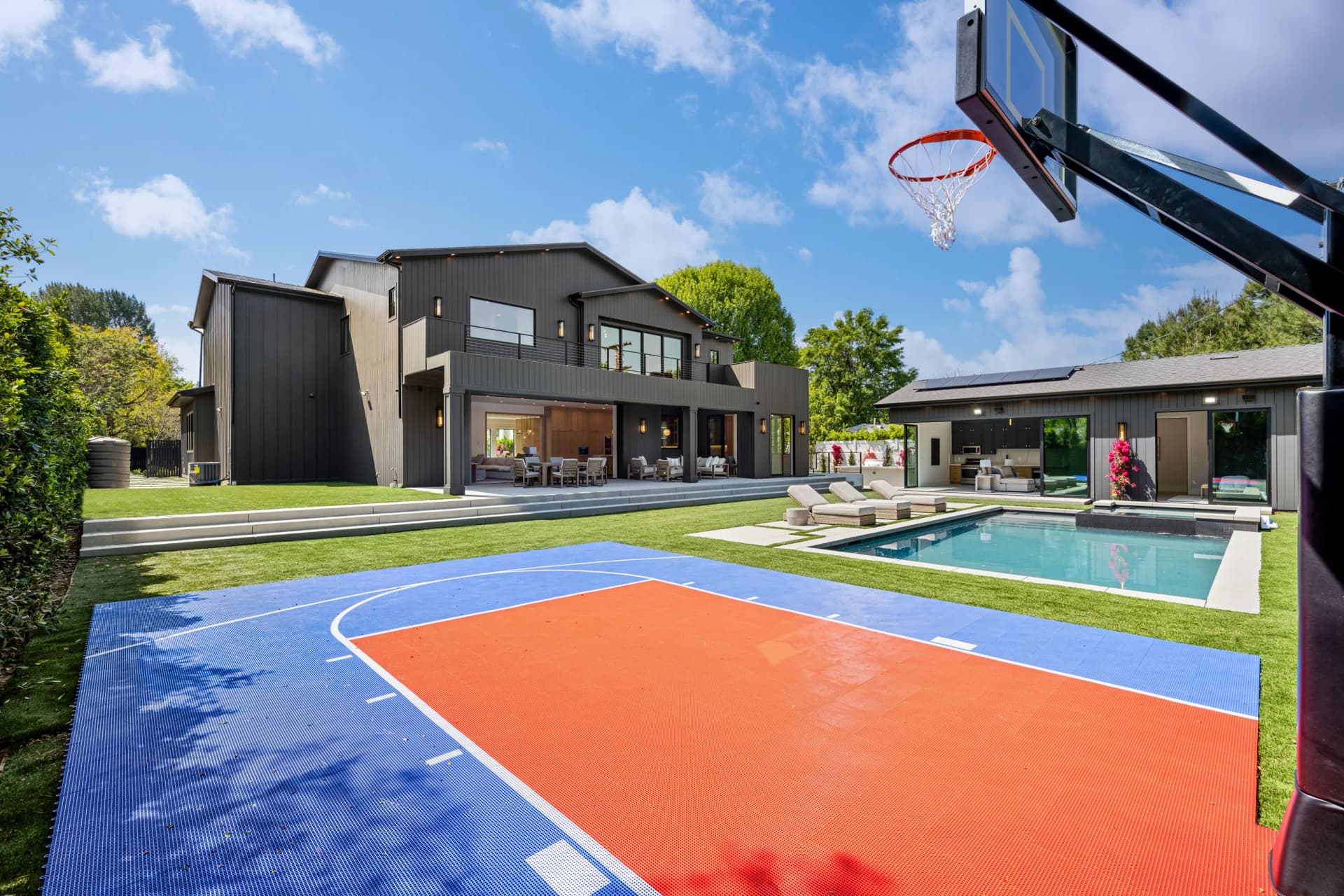 A modern dark grey house with a large backyard featuring a swimming pool, a patio with outdoor furniture, and a blue and orange basketball court.